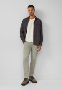 Schwarz-rote karierte Hemd, darunter ein weißes T-Shirt, hellgrüne Hose und braune Schnürstiefel. Lässiges, geschichtetes Outfit.