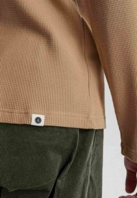 Beige, waffle-textúrájú, hosszú ujjú póló, kis logó címkével. Sötétzöld bordás nadrággal párosítva, hangsúlyozva a szövet kontrasztját és a színkombinációt.