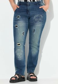 Jeans in denim di medio blu con un design distressed caratterizzato da strappi e un motivo di teschi arricchito con borchie. Fit dritto.
