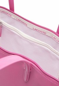 Sac fourre-tout rose avec une surface lisse, comportant une poche intérieure zippée blanche. Logo "LACOSTE" en rose clair le long de l'encadrement intérieur.
