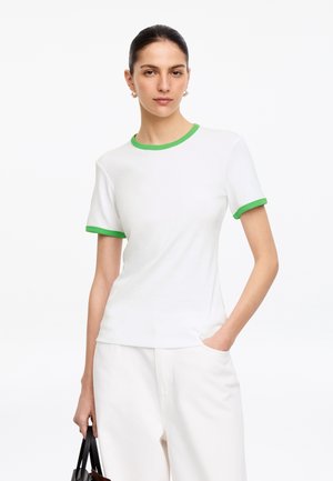 Femme portant une chemise blanche à manches courtes avec une bordure verte, pantalon blanc, tenant un sac noir avec une main dans la poche, expression neutre.