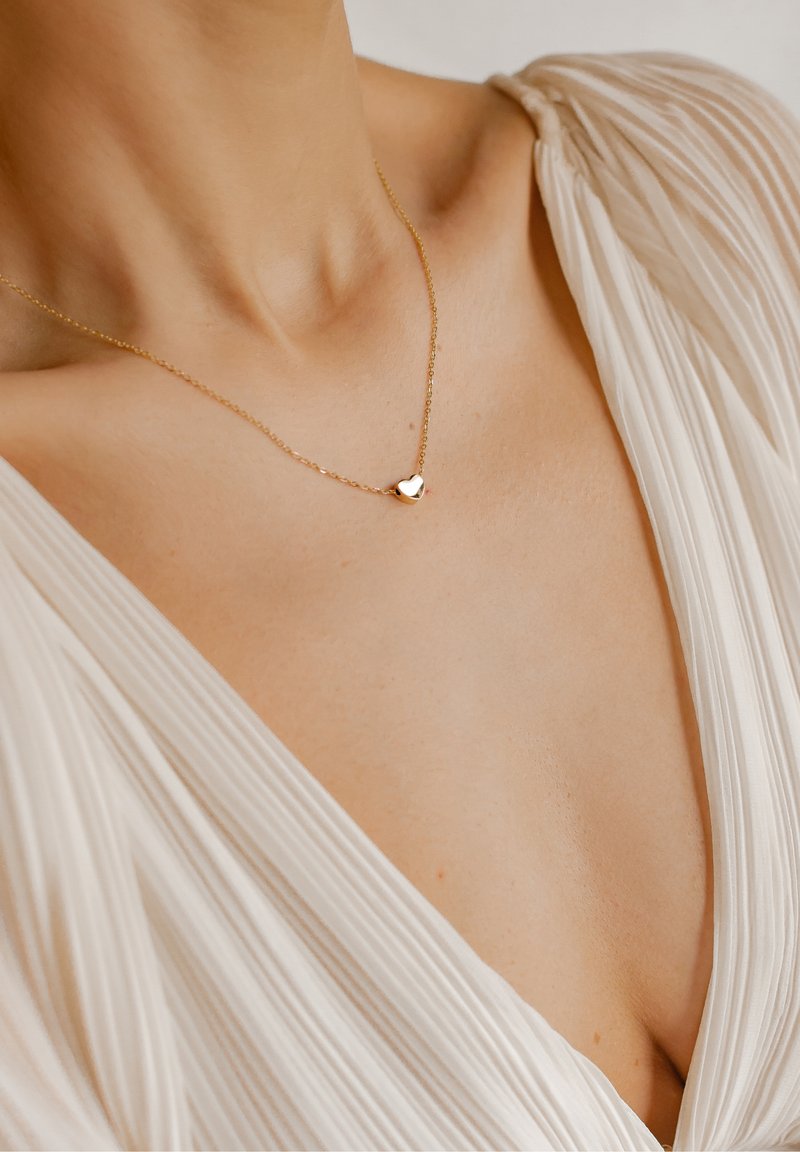Gros plan d'une personne portant un délicat collier en or avec un petit pendentif en forme de cœur et un vêtement blanc plissé à col en V.
