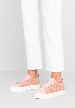 Persona che indossa pantaloni bianchi corti e sneaker rosa chiaro traforate con suole bianche spesse, in piedi su un pavimento grigio.