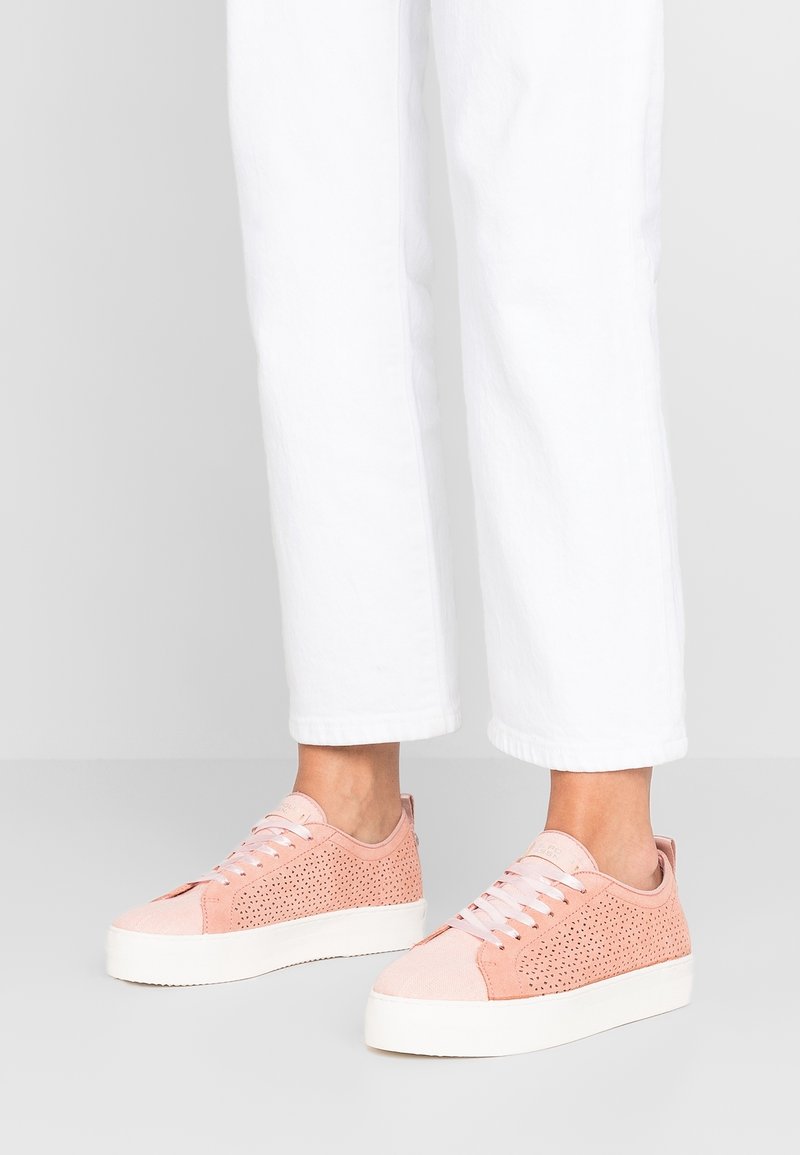 Personne portant un pantalon blanc court et des baskets rose clair perforées avec des semelles épaisses blanches, debout sur un sol gris.