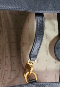 Schwarzer strukturierter Innenraum einer Tasche mit beige gefüttertem Innenfutter, das ein wiederholendes "GUESS"-Muster aufweist. Sichtbare goldene Metallakzente.