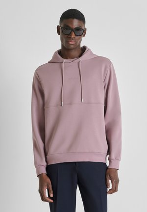 Man die zwarte zonnebril, een mauve hoodie met trekkoorden en donkere broek draagt, staand tegen een effen lichtgrijze achtergrond.