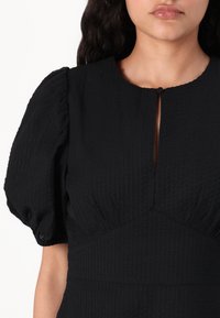 Czarna sukienka z fakturowaną, marszczoną tkaniną, krótkimi bufkami, okrągłym dekoltem z guzikiem i niewielkim rozcięciem z przodu.