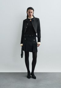 Svart kort jacka med guldknappar, texturblus i svart med volanger, svart plisserad kjol, ogenomskinliga strumpbyxor och svarta skor. Liten handväska.