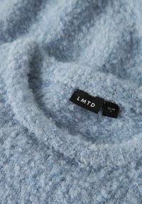 Gaiši zils teksturēts apģērba gabals ar mīkstu, pūkainu virsmu. Tam ir uzlīme ar uzrakstu "LMTD" un izmēra informāciju.