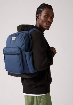 Zaino in tessuto blu con più scomparti, incluso una tasca anteriore con zip e una tasca laterale in rete, indossato sulla schiena di un modello.