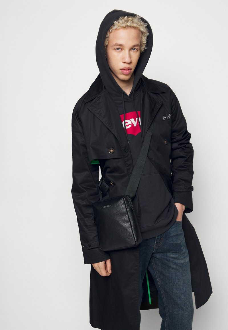 Cappotto nero sopra un felpa scura con logo rosso, jeans blu e una borsa a tracolla in pelle nera con logo impressionato.