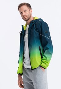 Huvtröja med en ombre-gradient från mörkblå till klargrön, tillverkad av lätt material, med dragkedjefickor och kontrasterande neon-accenter.