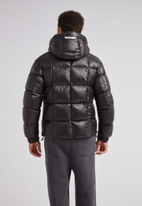 Giacca a vento nera con design trapuntato, finitura lucida, cappuccio e polsini elasticizzati. Indossata sopra pantaloni grigi, evidenziando una silhouette slanciata.