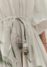 White, lightweight dress with floral green patterns, cinched waist, and decorative tassels in beige, green, and blue. Textured fabric details.