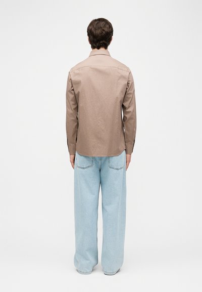 Άτομο με την πλάτη, φοράει μπεζ μακρυμάνικο πουκάμισο και χαλαρά ανοιχτόμπλε τζιν, σε απλό λευκό φόντο.
