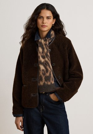 Femme aux longs cheveux foncés portant une veste en laine marron foncé boutonnée, une écharpe à motif léopard et un jean bleu foncé avec une main dans la poche.