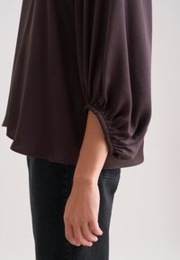 Pullover marrone scuro con maniche elastiche arricciate. Il tessuto appare morbido con una superficie liscia. Indossato con jeans scuri.