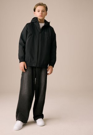 Tânăr purtând o jachetă neagră cu glugă și fermoar, blugi largi închiși la culoare și adidași albi, stând în fața unui fundal bej simplu.