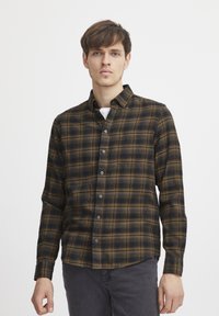 Camisa de franela a cuadros en marrón y negro, con cierre de botones, mangas largas y un bolsillo en el pecho, llevada sobre una camiseta blanca.