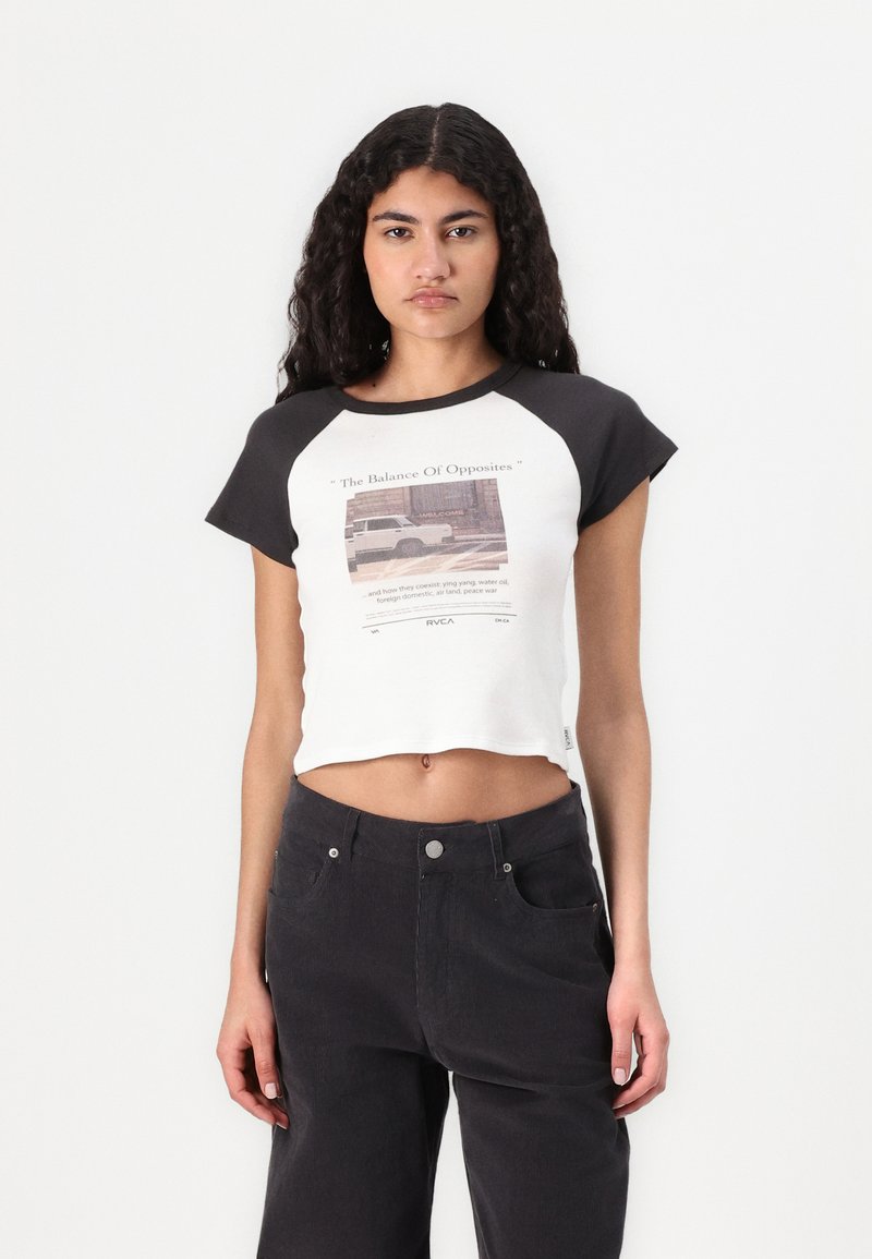 Top corto de algodón con mangas raglán negras, que presenta un diseño gráfico impreso y texto en la parte delantera. Combinado con pantalones negros de cintura alta.