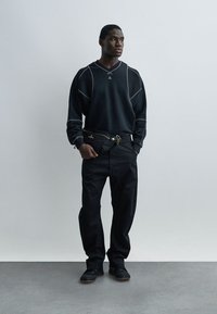 Άνδρας στέκεται μπροστά σε απλό τοίχο φορώντας μαύρο φούτερ με λευκές ραφές, μαύρο παντελόνι, μαύρα αθλητικά παπούτσια και μαύρη τσάντα ώμου.