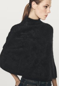 Kvinna iklädd en svart fluffig poncho med hög krage och blå jeans, som tittar åt höger mot en neutral bakgrund.