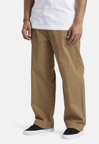 Kaki vidde bukser lavet af bomuldsblandingsstof, med en glat tekstur og sidelommer, parret med sorte sneakers og en hvid t-shirt.