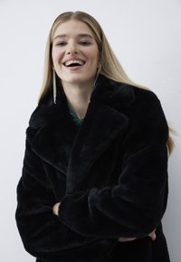 Vrouw met een glimlach en lang blond haar, gekleed in een zwarte faux bontjas, een groene top en lange hangende oorbellen met strassstenen, tegen een witte achtergrond.