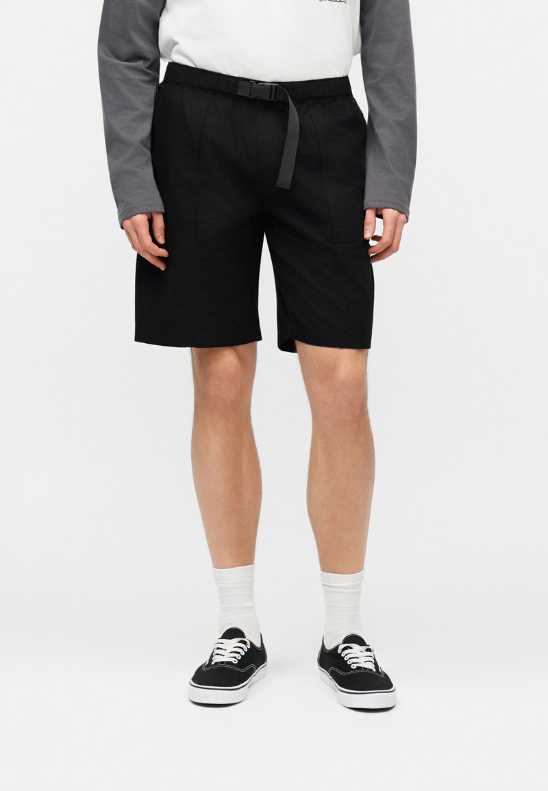 Άτομο που φοράει μαύρα σορτς μέχρι το γόνατο με ρυθμιζόμενο ζωνάκι, λευκές κάλτσες και μαύρα αθλητικά παπούτσια χαμηλής κοπής, στέκεται μπροστά σε απλό φόντο.