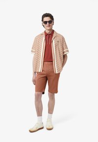Camicetta a maniche corte con motivo geometrico arancione e bianco, sovrapposta a un polo rosso, abbinata a pantaloni corti ruggine e sneakers crema.
