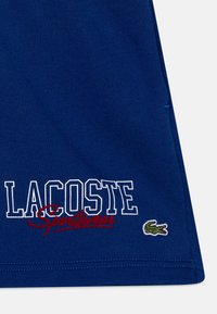 Modré bavlněné tričko s nápisem "LACOSTE" v bílé barvě a "Sportwear" v červené barvě, s malým zeleným krokodýlem v pravém dolním rohu.