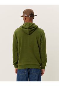 Felpa verde oliva con coulisse, tessuto morbido, polsini e orlo a coste; indossata con un berretto marrone. Nessuna vista frontale visibile.