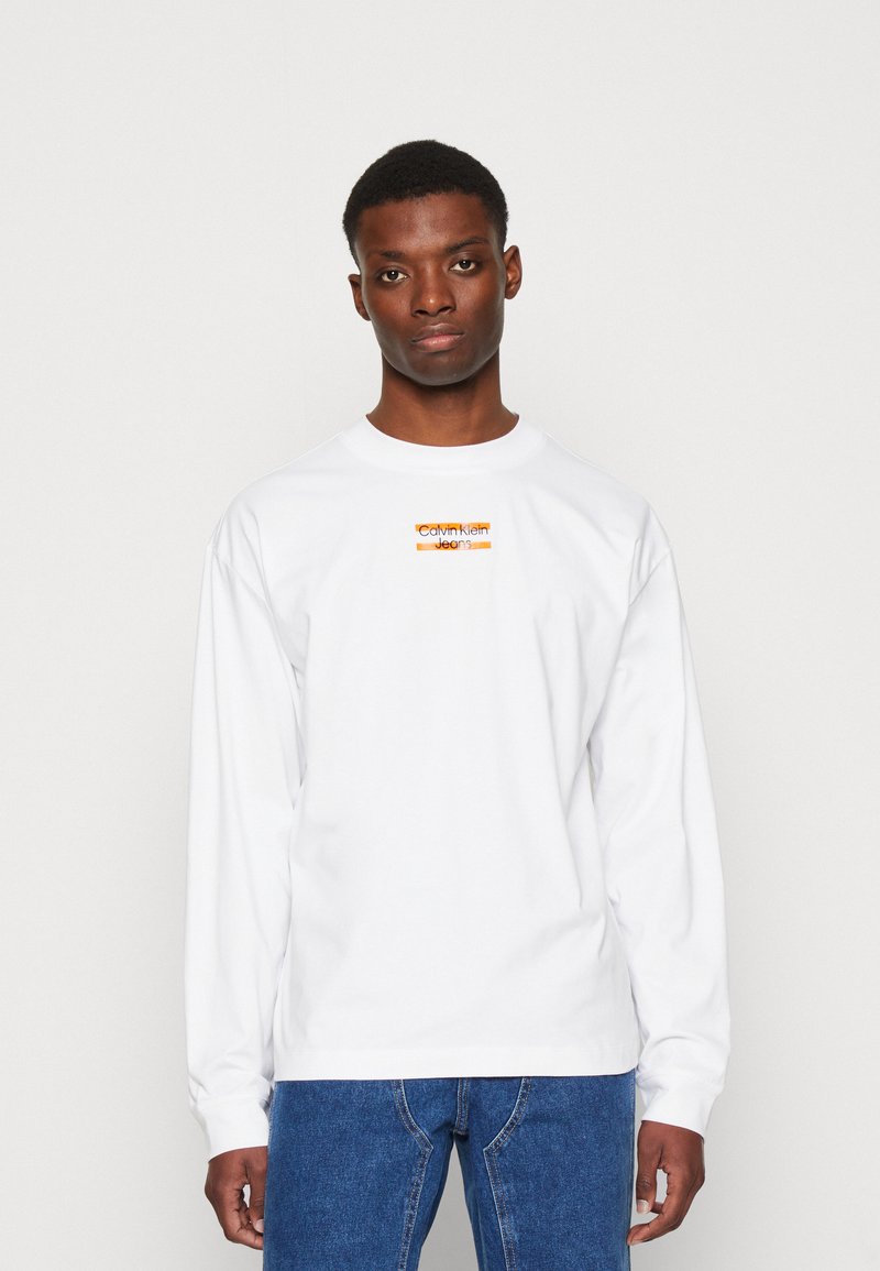 Fehér hosszú ujjú póló, amelyen egy kis téglalap alakú narancssárga logo található a mellrészen, rajta a "Calvin Klein Jeans" szöveggel. Normál szabású, pamut anyagból készült.