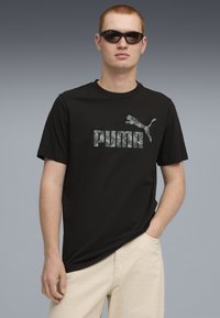 Sort kortærmet t-shirt med et fremtrædende gråt "PUMA" logo og en springende puma grafik. Modellen har solbriller på og bærer cremefarvede bukser.