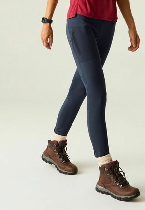 Personne portant un pantalon bleu foncé, des bottes de randonnée marron avec des chaussettes beige et une chemise rouge, marchant, avec une montre noire au poignet gauche.