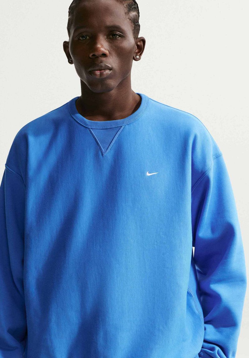 Junger schwarzer Mann in blauem Nike-Sweatshirt, steht vor schlichtem weißem Hintergrund und blickt mit neutralem Ausdruck geradeaus.