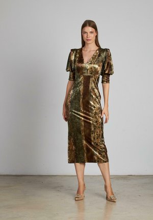 Mujer de pie con un vestido de terciopelo verde y marrón de longitud media, con mangas acampanadas y tacones beige, sobre un fondo liso.