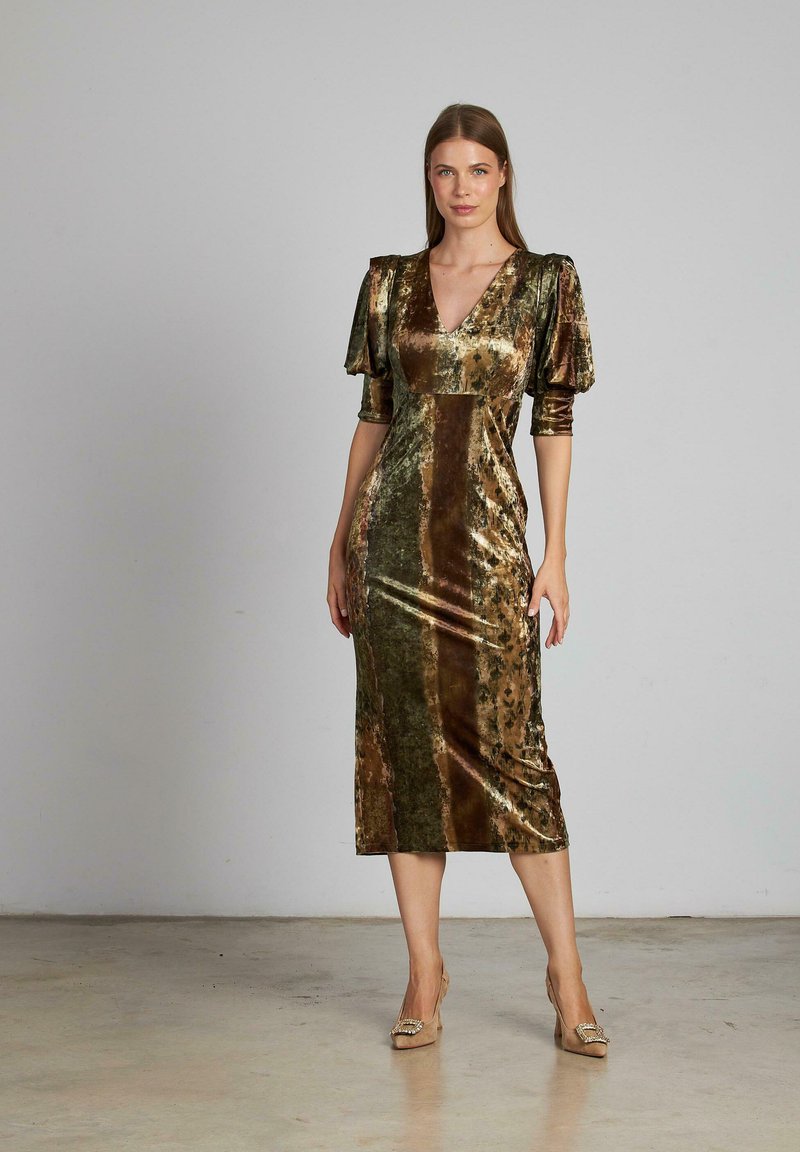 Donna in un abito di velluto verde e marrone a media lunghezza con maniche a sbuffo e tacchi beige su uno sfondo semplice.