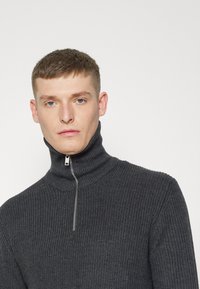 Mørkegrå, ribbet strikkegenser med høy krage og halv glidelås foran. Stoffet ser mykt og teksturert ut. EnkeltDesign uten fremtredende detaljer.