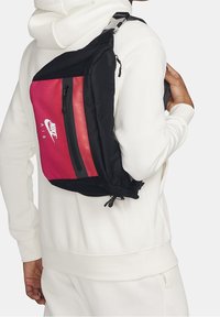 Nike Air olkalaukku, jossa on punainen ja musta muotoilu. Suuri vetoketjullinen tasku, sileä materiaali ja säädettävä hihna.