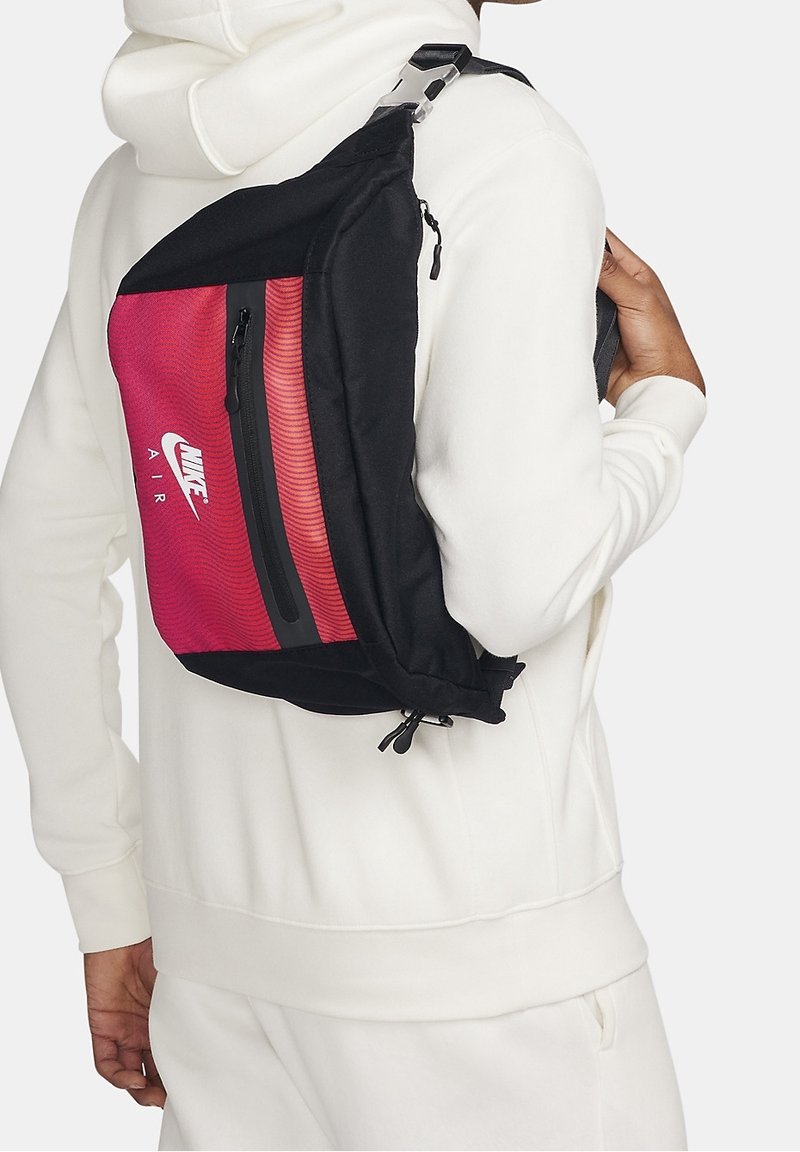 Nike Air olkalaukku, jossa on punainen ja musta muotoilu. Suuri vetoketjullinen tasku, sileä materiaali ja säädettävä hihna.