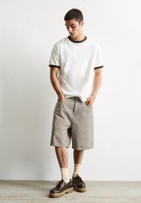 T-shirt en coton blanc avec des accents marron, shorts beiges et chaussures à lacets marron avec une semelle épaisse. Le modèle se tient les mains dans les poches.