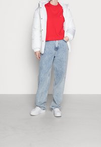 Veste matelassée blanche, chemise rouge à manches courtes, jean bleu clair ample et baskets blanches. Ensemble décontracté avec des contrastes de couleurs distincts.