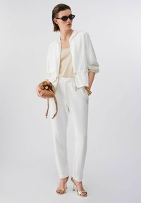 Femme portant une veste blanche à fermeture éclair, un haut crème, un pantalon blanc à cordon, des talons dorés, des lunettes de soleil, des bracelets dorés, tenant une pochette en cuir marron.