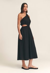Vestido maxi negro de un solo hombro con diseño recortado en la cintura. Tela suave, ajuste suelto y cintura elástica. Combinado con sandalias minimalistas.