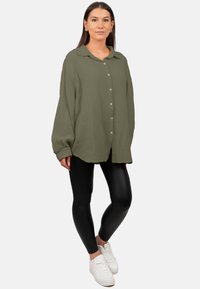 Olijfgroene, oversized knoopshirt met een gestructureerde stof, lange mouwen en een ronde zoom, gecombineerd met zwarte leggings en witte sneakers.