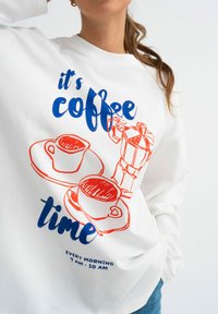 Biała bluza z niebieskim i czerwonym graficznym nadrukiem filiżanek kawy oraz ekspresu do kawy, z tekstem "czas na kawę" oraz "KAŻDE RANO 7:00 - 10:00."
