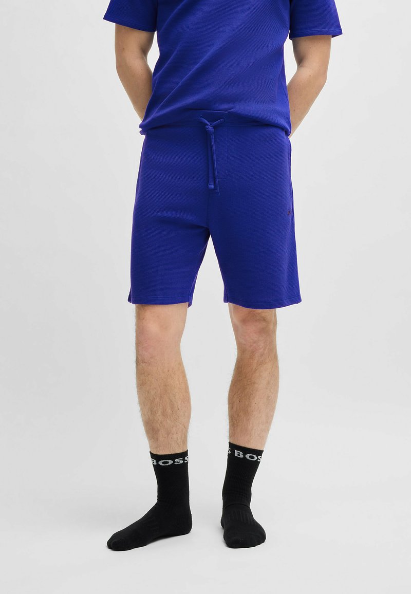 Blauwe sportshorts van een textiel met structuur en een verstelbare trekkoordtaille. Combinatie met zwarte sokken met een wit logo.