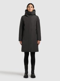 Ilga juoda žieminė striukė su aukštu apykakliu ir gaubtu, turinti priekinį užtrauktuką, šonines kišenes ir aptakų siluetą. Dėvima su juodomis tamprėmis ir batais.