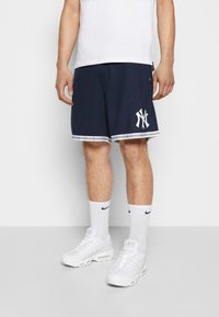 Shorts sportifs bleu marine avec logo blanc ; bordure blanche contrastante à l'ourlet. Portés avec des chaussettes blanches et des baskets.